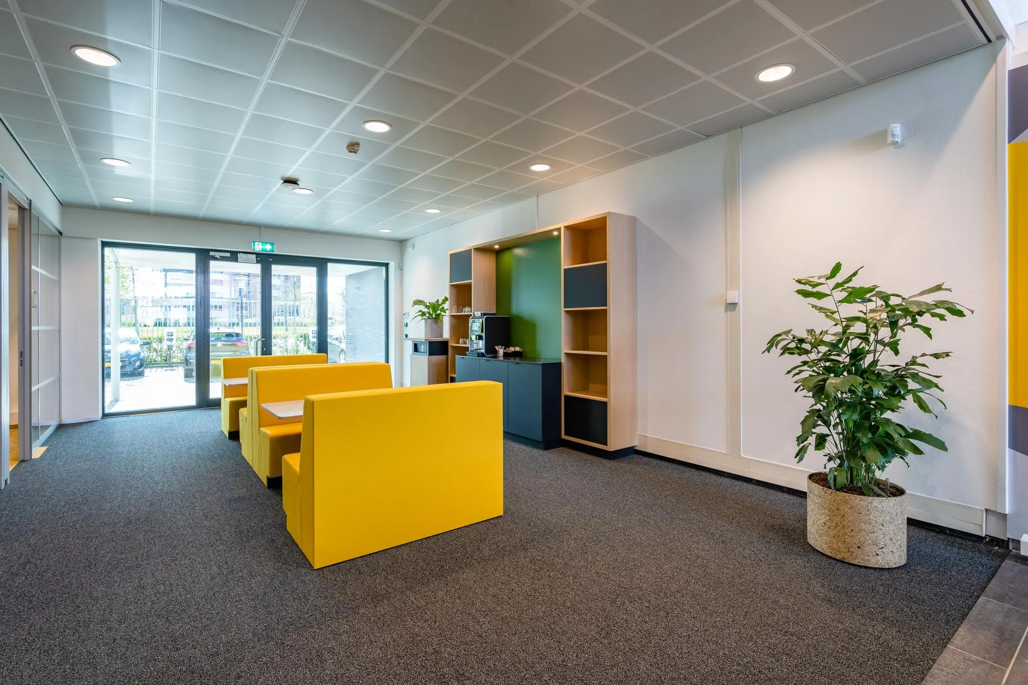 Moderne wachtruimte aan de Fascinatio Boulevard met felgele zitbanken, een koffiemeubel en een grote plant in een pot.